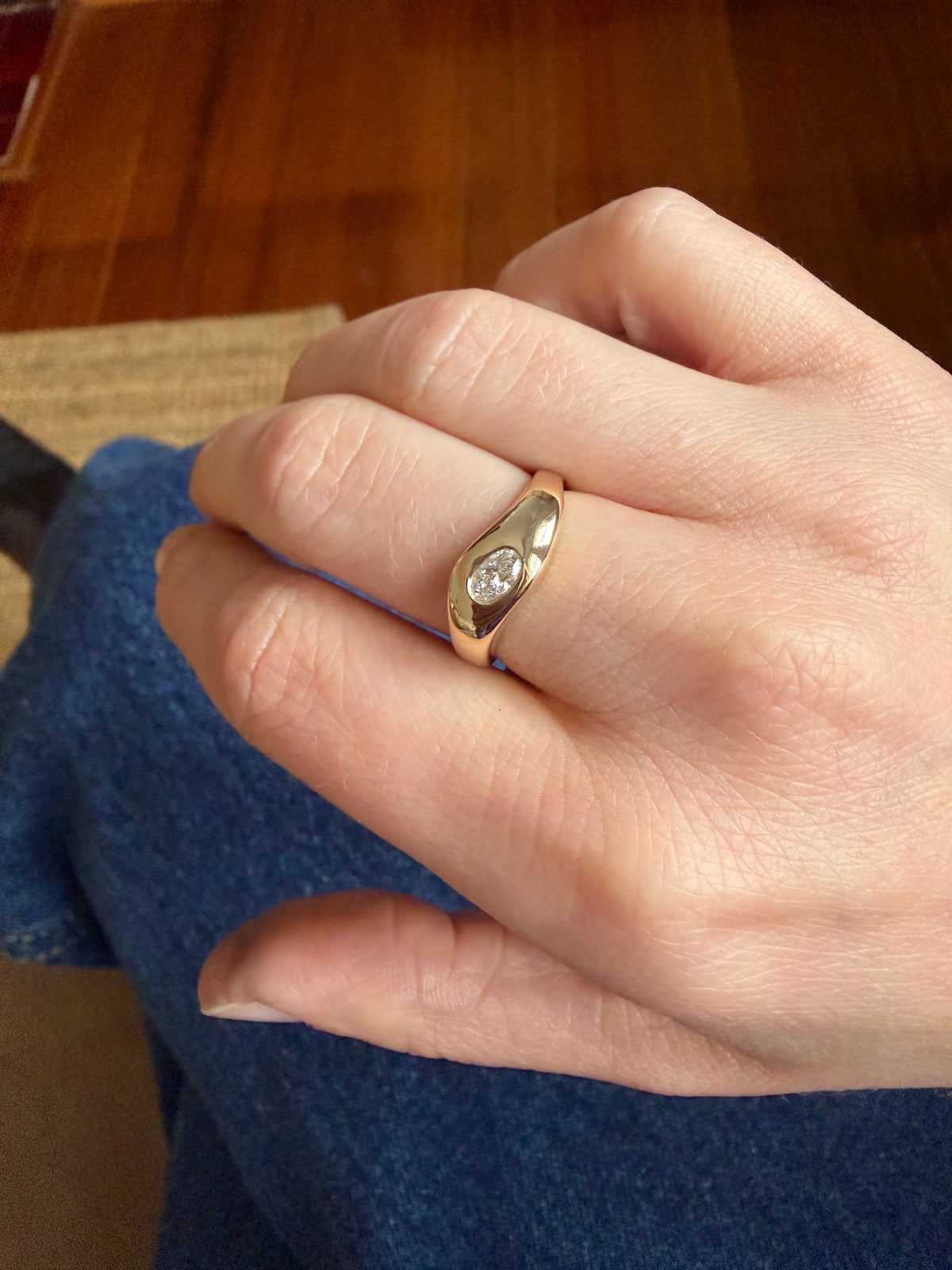 A close-up of a person's hand wearing a polished yellow gold ring with a single round-cut clear diamond, set against a background of blue clothing and a wooden floor.