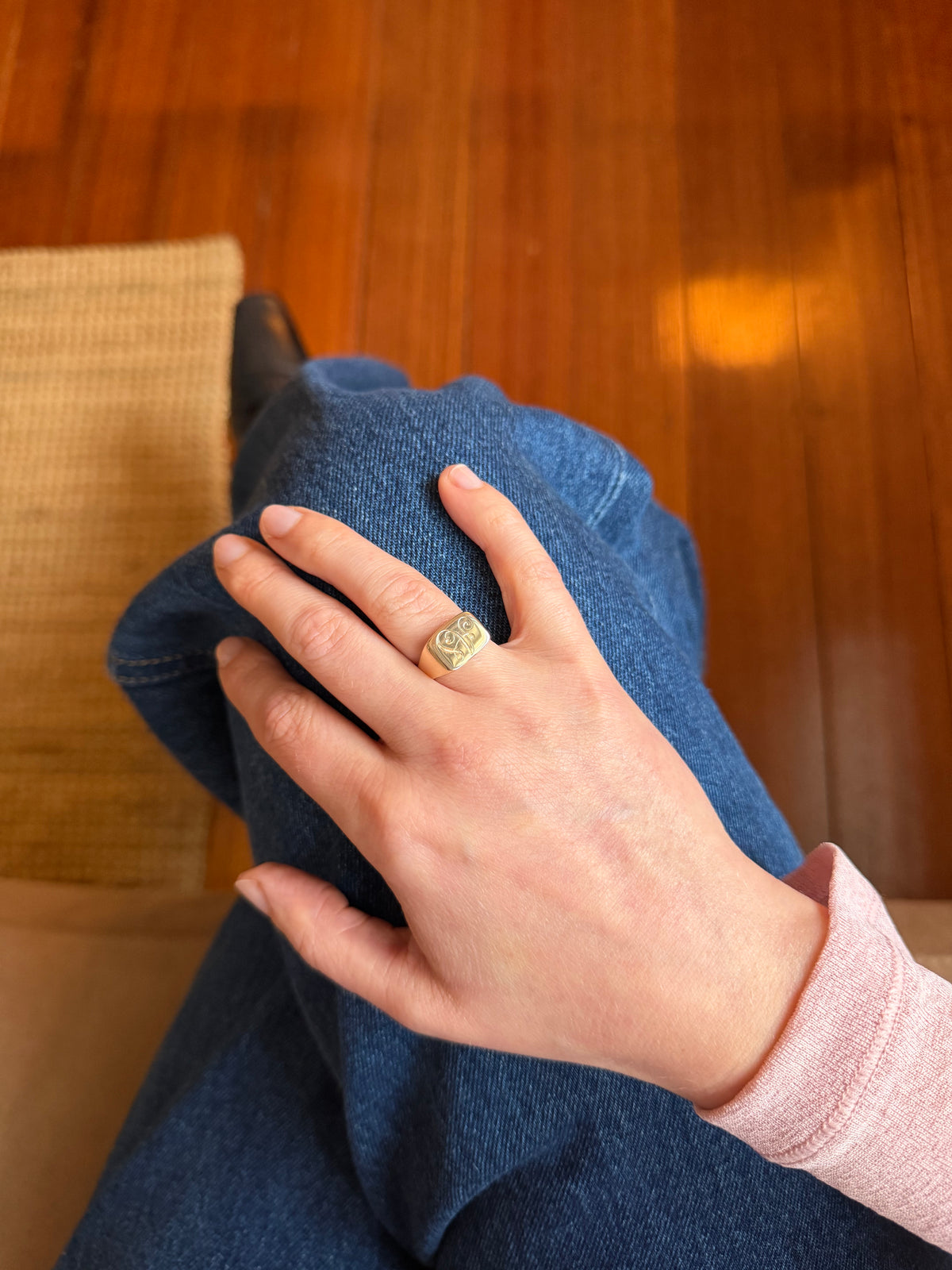 A close-up lifestyle shot of a person's hand resting on their lap, wearing a textured gold signet ring. The person is wearing blue jeans and a light pink long-sleeve top, with a warm wooden floor visible in the background.