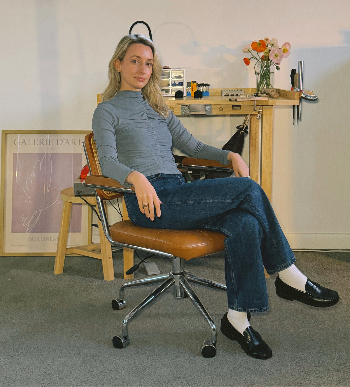 Aisling sits in a brown leather office chair with a chrome base in a styled room. She is wearing a gray ribbed mock neck top, blue jeans, white socks, and black loafers.