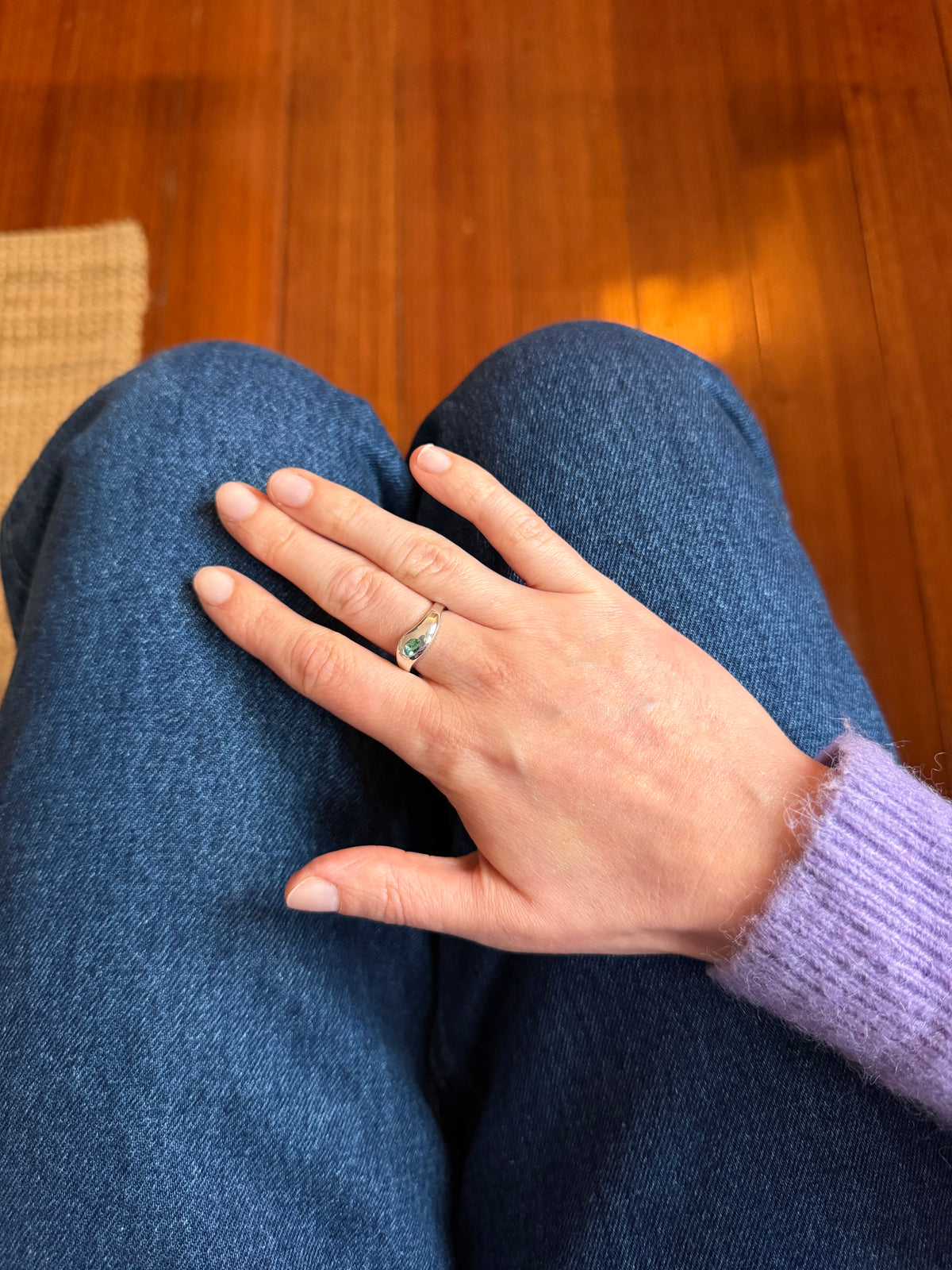 A silver rounded ring on a persons hand, resting on blue jeans with a light purple sweater