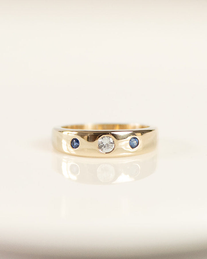A gold band ring with a central clear round gemstone and two small blue round gemstones on either side, resting on a white reflective surface.
