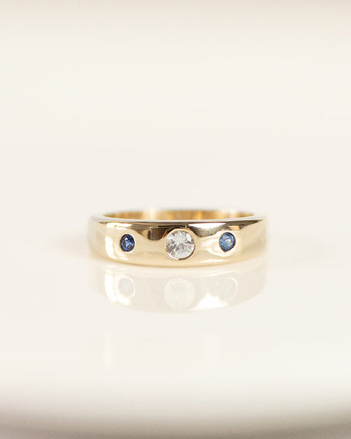 A gold band ring with a central clear round gemstone and two small blue round gemstones on either side, resting on a white reflective surface.