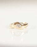 A gold ring with a small clear white diamond, resting on a white reflective surface.