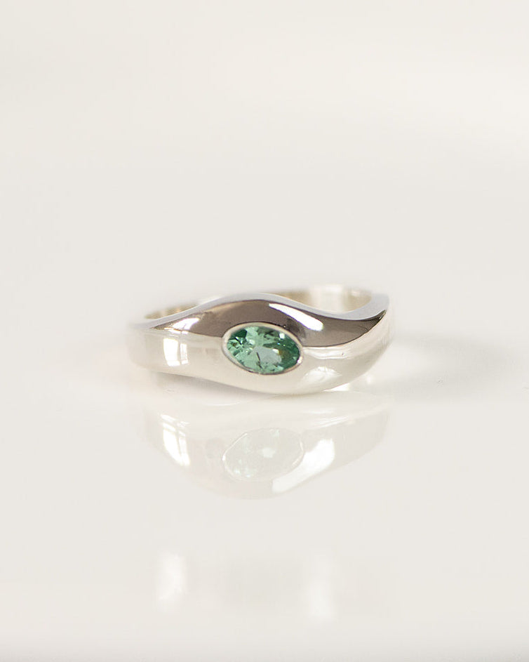A silver ring with a small oval green tourmaline, displayed on a white reflective surface.