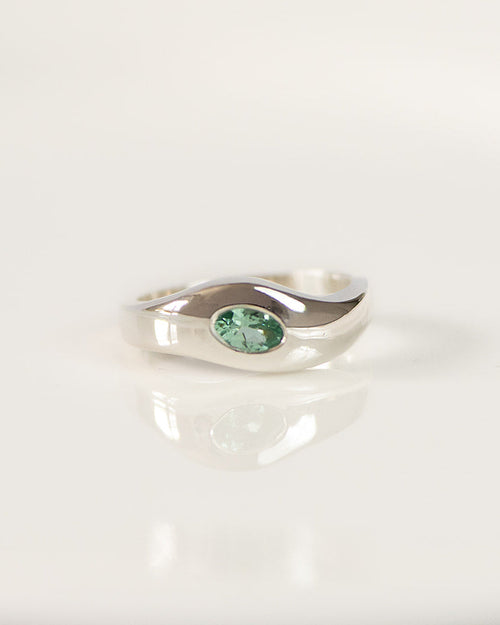 A silver ring with a small oval green tourmaline, displayed on a white reflective surface.