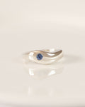 A silver wave-shaped ring with a small round blue sapphire, resting on a white ceramic plate.