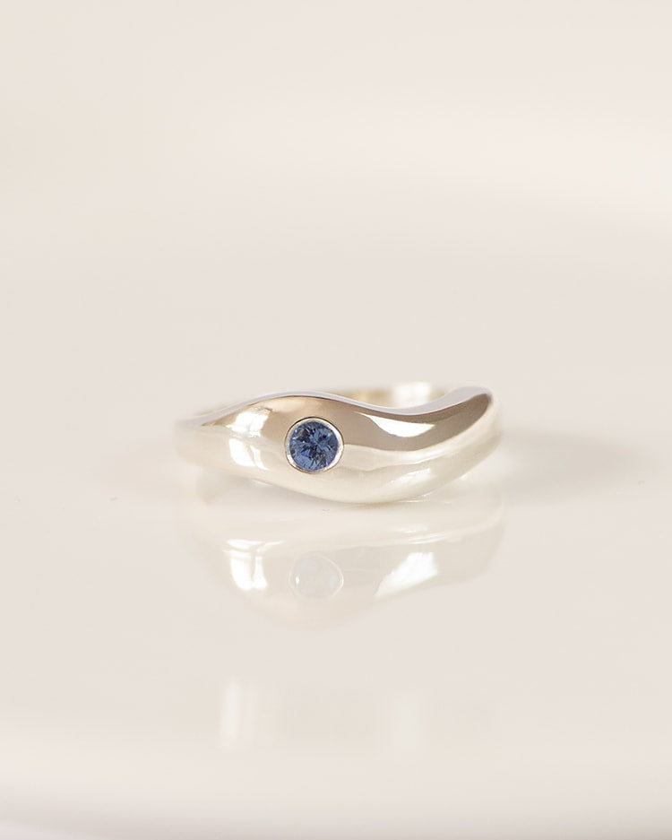 A silver wave-shaped ring with a small round blue sapphire, resting on a white ceramic plate.