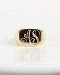 A gold signet ring featuring the intertwined initials 'AS' in polished gold, displayed on a white ceramic plate.