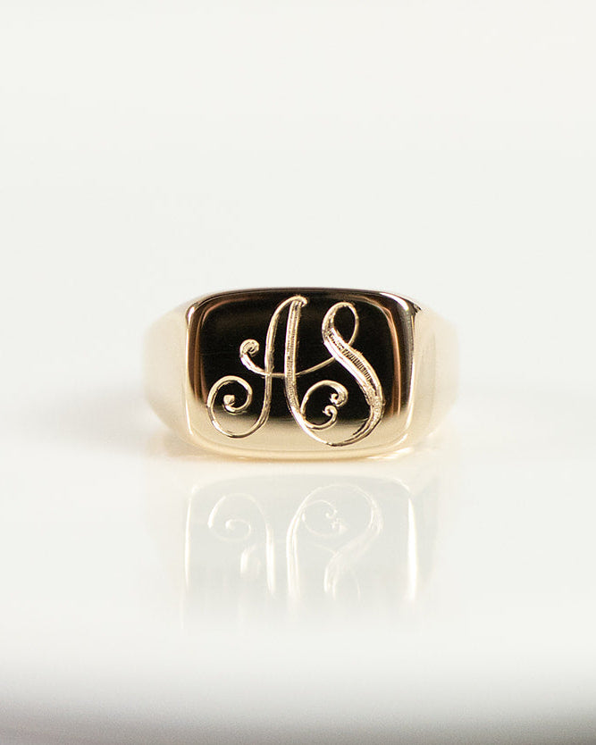A gold signet ring featuring the intertwined initials 'AS' in polished gold, displayed on a white ceramic plate.