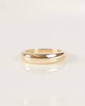 A minimalist gold band ring with a smooth, polished finish, resting on a clean white surface with a soft reflection.
