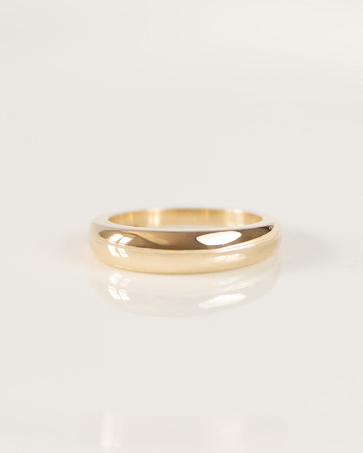 A minimalist gold band ring with a smooth, polished finish, resting on a clean white surface with a soft reflection.