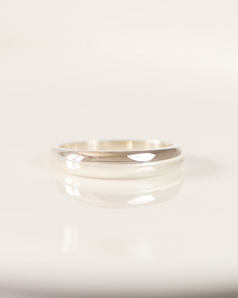 A minimalist polished silver band ring resting on a white ceramic plate with a soft reflection.