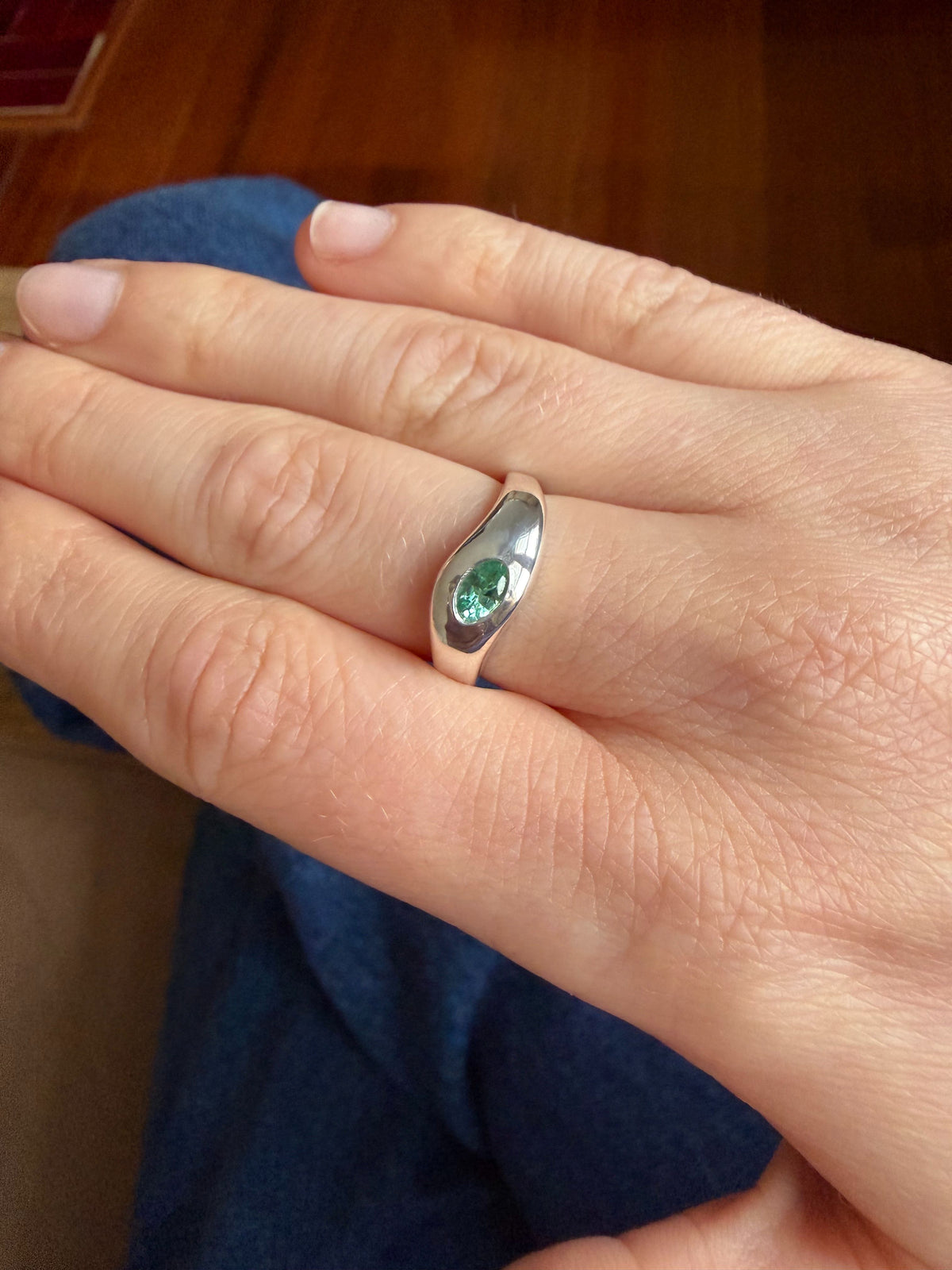A silver ring with a small green oval stone, worn on a person's middle finger against a background of blue jeans and a wooden floor.