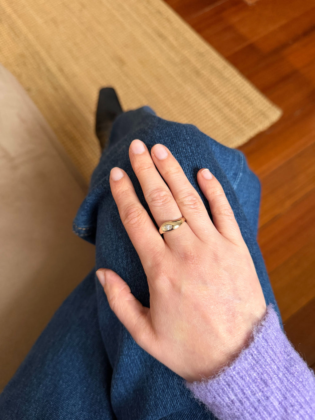 A gold band ring with a small white diamond set on a person's ring finger, shown while wearing blue jeans and a lavender sweater in an indoor setting.