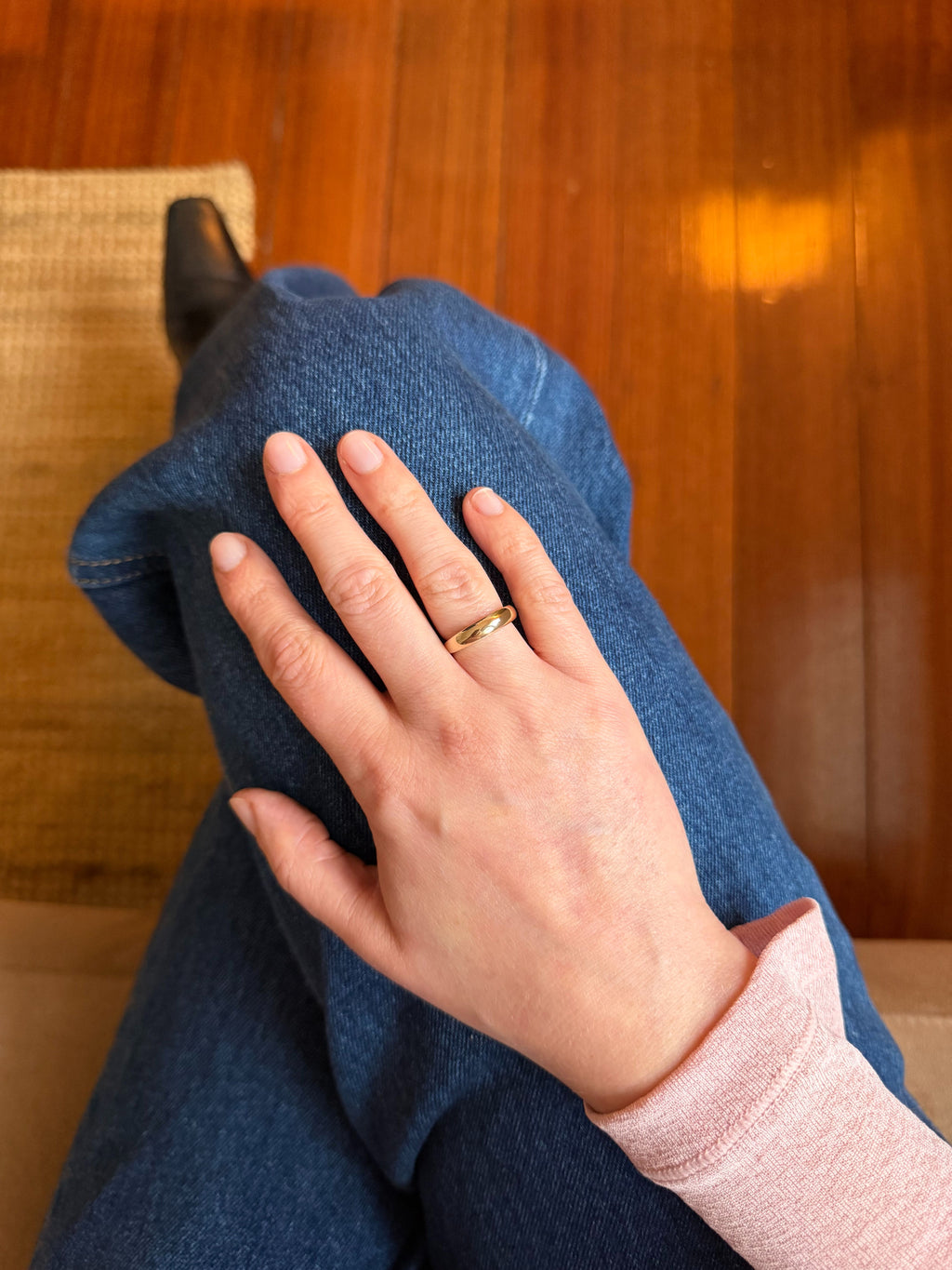 A close-up of a hand resting on blue jeans, wearing a simple gold band ring. The person is also wearing a light pink long-sleeve top, and the background shows a warm wooden floor.