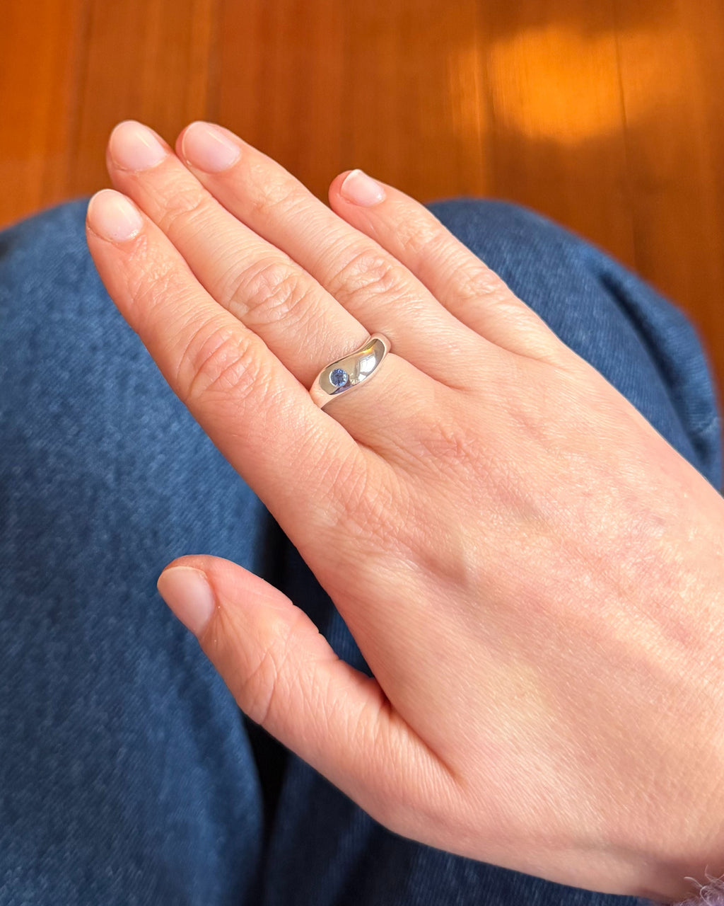 A silver band ring with a small oval blue sapphire is worn on the ring finger of a person's hand, shown in a close-up shot against a background of blue jeans and a wooden floor.