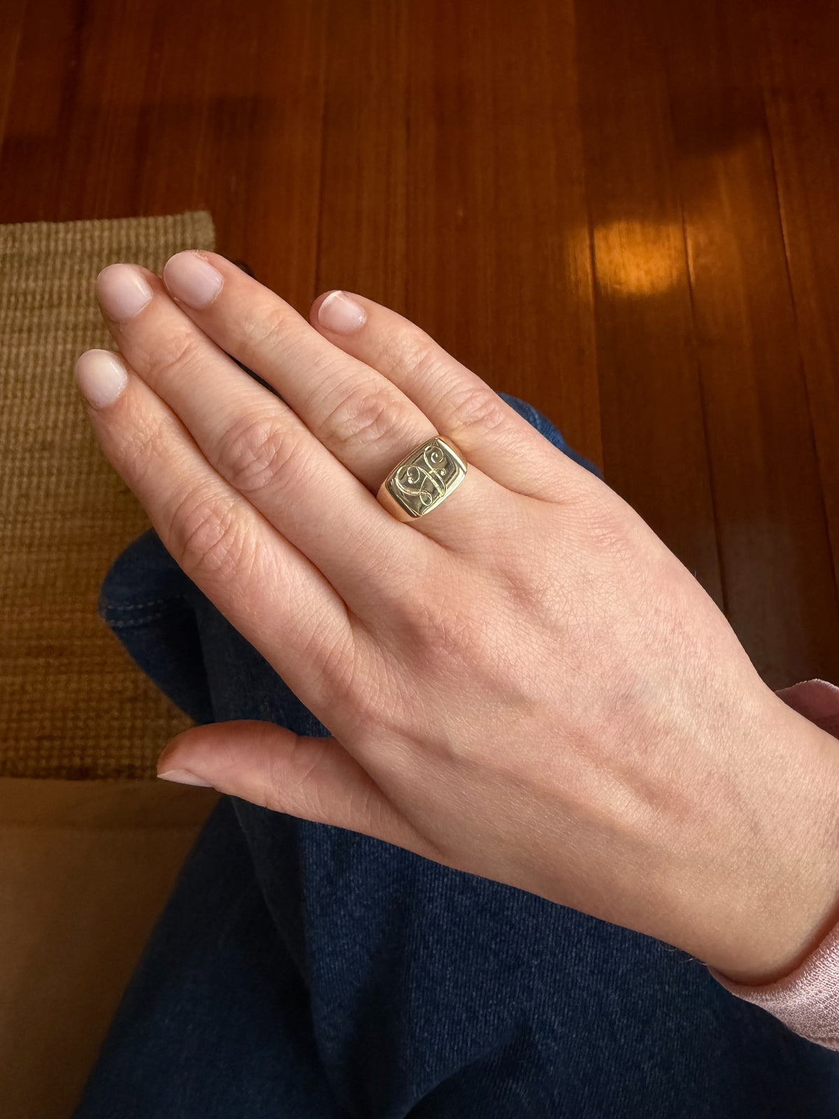 A gold signet ring with a raised circular emblem on the side, worn on a person's ring finger. The hand is resting on dark blue denim, with a wooden floor and a woven mat visible in the background.