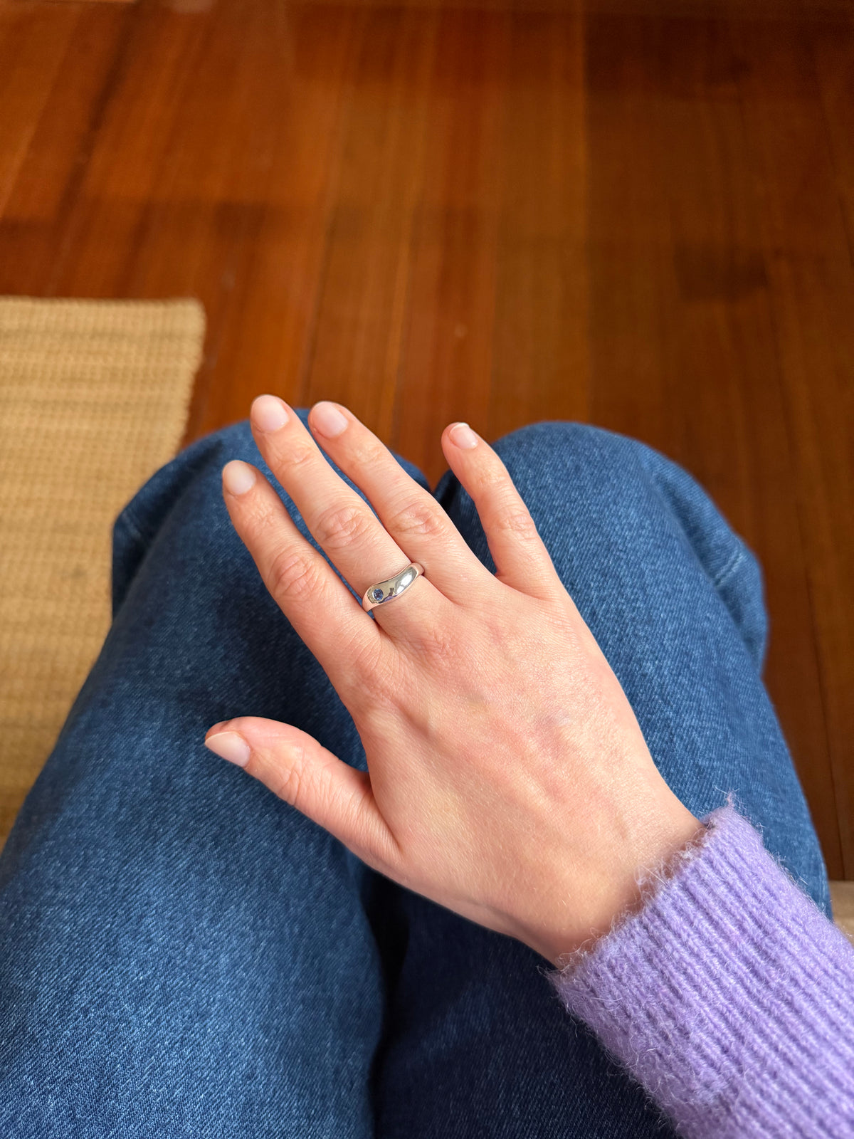 A silver band ring with a small blue stone is worn on the ring finger of a person's hand, which is resting on their lap. The person is wearing blue jeans and a light purple knit sweater, with a wooden floor and woven rug in the background.