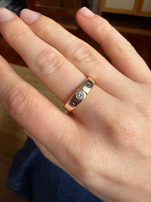 A close-up of a hand wearing a gold band ring with a central round diamond and two small round blue sapphires, set against an indoor background.